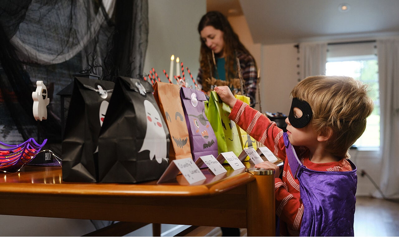 mother setting up halloween decorations while child in costume looks at halloween themed treat bags