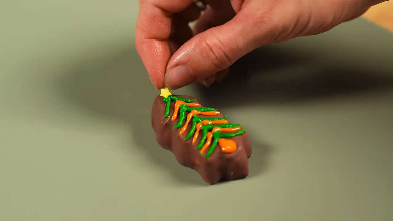 hand placing a yellow star sprinkle on a reeses tree decorated with green and orange icing