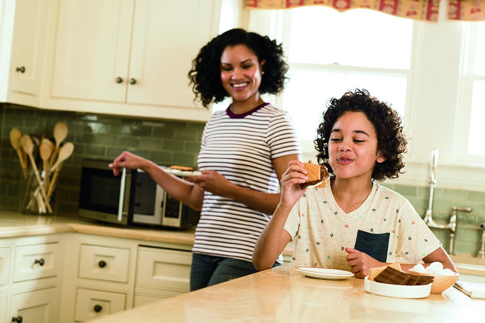 Child eating a S'more at the table with parent standing next to them