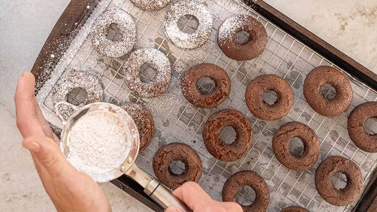 sprinkling powdered sugar over doughnuts on wire cooling rack
