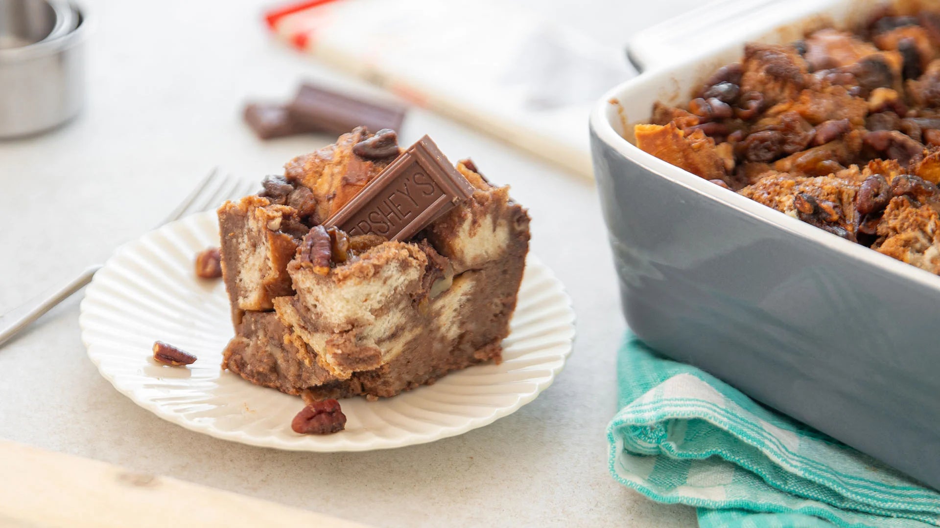 slice of chocolate bread pudding with pecans and a milk chocolate piece on a plate beside the casserole dish