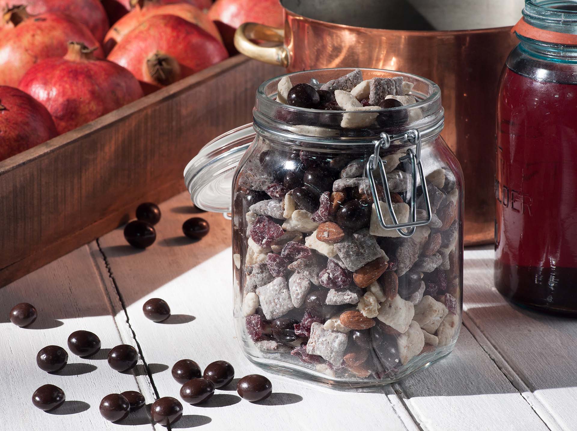 Checkered BROOKSIDE Snack Mix in a large glass jar on a table with Brookside dark chocolate pieces scattered in front of the jar. Behind the jar are various pots and pomegranates in a crate. 