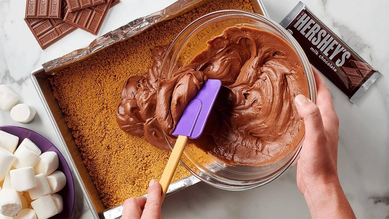 person spreading brownie mixture on top of a graham cracker crust
