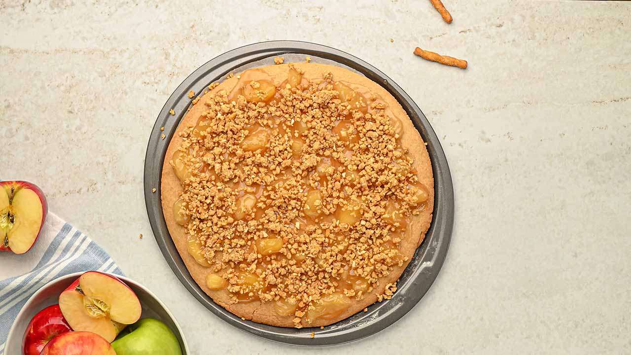 removing the cookie crust from the oven and topping with apple filling and pretzel crumble