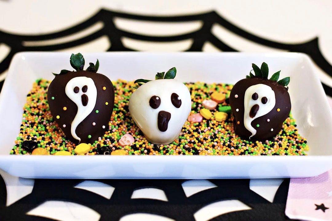 three chocolate covered strawberries decorated as ghosts on a tray of sprinkles